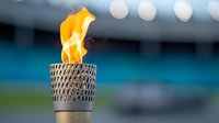 Burning ceremonial torch with bright orange flame against blurred stadium background, metallic decorative holder with geometric pattern design creates dramatic atmosphere.