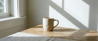 Ceramic mug on wooden table by window with natural sunlight casting shadows, minimalist morning coffee scene for lifestyle and home decor projects.