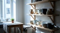Wooden shelves displaying ceramic bowls and dishes in neutral tones, with a small potted plant on a wooden table by a bright window, creating a minimalist home decor and storage solution.