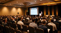 Business conference presentation with speaker addressing large audience in modern hotel ballroom with professional lighting and projection screen.