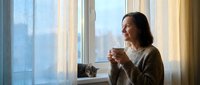 Young Asian woman enjoying morning coffee by window with natural light streaming through curtains creating peaceful domestic atmosphere for lifestyle content.