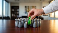 Male hand in white business shirt selecting green pawn from group of gray pawns on wooden table, representing leadership, uniqueness, recruitment, and business strategy concepts.