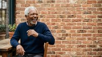 Senior African American man with white hair, wearing navy blue sweater, sitting outdoors against brick wall. Genuine smile, relaxed posture, lifestyle portrait for aging, retirement, wellness.