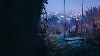 Wooden swing hanging from tree branches in twilight garden with blurred city lights creating dreamy bokeh background atmosphere for relaxation and solitude.