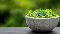 Fresh homemade guacamole with cilantro and avocado served in stone bowl on wooden table against blurred green garden background.