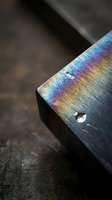 Iridescent rainbow colors on dark metal surface with water droplets creating prismatic light reflection and optical interference patterns.