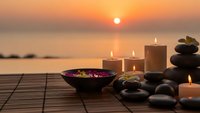 Spa wellness setup with candles, stones and flowers at sunset by ocean for relaxation and meditation therapy treatments.