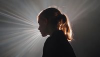 Dramatic silhouette of young woman with ponytail against bright rays of light creating ethereal atmosphere and spiritual mood in dark setting.
