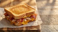 Golden grilled cheese sandwich with crispy bacon on parchment paper over rustic wooden cutting board. Comfort food breakfast or lunch meal ready to eat.