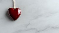 Red glossy heart pendant hanging on string against white marble background, minimalist romantic composition with copy space for valentines day messages and designs.