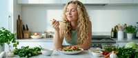 Young blonde Caucasian woman enjoying healthy salad meal in modern kitchen with fresh vegetables and herbs on counter for nutritious eating lifestyle.