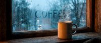 Steaming mug on a wooden windowsill with coffee written on a foggy rain-covered window, creating a cozy autumn atmosphere.