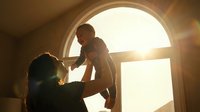 Mother holding up baby in warm sunlit room with arched window creating intimate bonding moment during golden hour light streaming through glass.