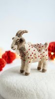 Felted goat wearing festive red berry patterned sweater, standing on white surface with blurred red ornaments in background. Handmade wool craft decoration for holiday seasonal displays and celebrations.