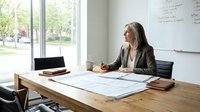 Mature woman with long dark hair and light skin tone, wearing business casual attire, reviewing architectural blueprints at wooden desk in bright office with whiteboard and window view.
