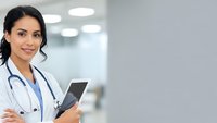 Confident female doctor with stethoscope holding tablet in modern hospital corridor, smiling at camera with arms crossed in professional healthcare setting.