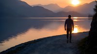 Silhouette of man walking along peaceful lake shore at golden sunset with mountain ranges reflecting in calm water creating serene outdoor adventure scene.