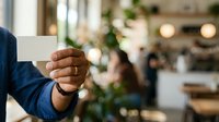 Man in blue shirt holding blank white business card at networking event with blurred indoor background and greenery, suitable for corporate branding and professional communication concepts.