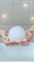 Female hands holding white foam soap bubble in bathroom with soft bokeh lights. Gentle skincare and relaxation concept for spa and wellness designs.
