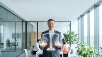 Professional businessman in suit holding digital tablet displaying holographic data charts in modern glass office environment for corporate presentations.