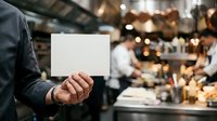 Caucasian male holding blank white card at social gathering with blurred guests and warm lighting in background, suitable for event invitations, announcements, promotions.