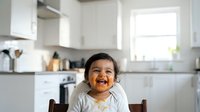 Young toddler with dark hair and medium skin tone sitting in kitchen, wearing light colored clothing, smiling at camera with food on face, domestic setting.