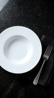 Empty white bowl with fork on black napkin against dark granite countertop, overhead view for restaurant, catering, and fine dining design projects.