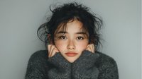 Young Asian woman in cozy gray sweater with turtleneck collar, messy hair and natural makeup, portrait against neutral background for lifestyle content.