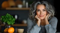 Confident mature woman with silver gray hair smiling at home. Natural beauty portrait with fresh basil plant and citrus fruit on kitchen counter in soft lighting.