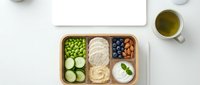 Healthy lunch box with compartments containing fresh vegetables, crackers, nuts and yogurt dip on white desk with tea cup and tablet for balanced nutrition.