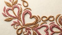 Handmade embroidery with pink and brown threads creating hearts and floral patterns on cream fabric background for craft projects.