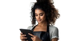 Young African American woman with curly hair wearing black apron using digital tablet, smiling while checking online orders or recipes against white background.