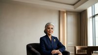 Mature woman with gray hair and glasses, wearing dark blazer with brooch, seated in modern office chair against neutral interior background with window.