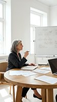 Mature woman with gray hair, Caucasian ethnicity, wearing dark blazer, sitting at wooden round table in bright office space, holding pen while reviewing whiteboard with flowchart diagrams, planning business strategy.