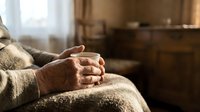 Elderly hands clasped together resting on lap in cozy living room setting with warm natural lighting and wooden furniture in background.