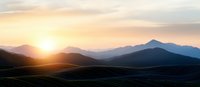 Panoramic mountain silhouettes against golden sunset sky, layered peaks fade into misty horizon creating atmospheric depth and tranquil wilderness scene.