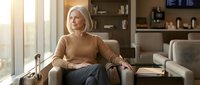 Senior Caucasian woman sitting comfortably in leather armchair at home, relaxing in modern living room with natural sunlight streaming through windows.