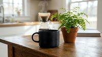 Cozy morning coffee setup with black mug and green plant on wooden table in bright kitchen interior with natural sunlight streaming through windows.