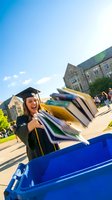 Excited young Caucasian female graduate in cap and gown tossing textbooks into a recycling bin on a sunny college campus.