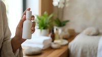 Person with dark skin tone holding white cosmetic bottle in bedroom with potted plants and neutral decor, suitable for beauty, skincare, wellness and home lifestyle marketing.