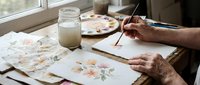 Senior artist painting delicate watercolor flowers at sunlit workspace with brushes, palette, and glass of water for creative art projects.