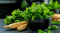 Fresh parsley leaves in black ceramic bowl with raw parsnip roots on dark wooden table, moody natural lighting for food photography and cooking blogs.