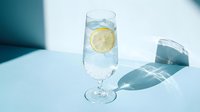 Glass of sparkling water with fresh lemon slice and ice cubes on light blue surface, casting shadows, suitable for beverage and wellness marketing.