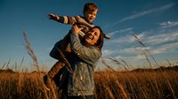 Happy mother carrying laughing son on shoulders in golden wheat field during sunset, family bonding moment in countryside nature landscape.