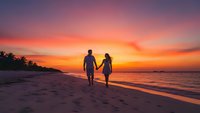 Romantic couple walking hand in hand on tropical beach at sunset with vibrant orange and pink sky reflecting on calm ocean waters during golden hour.