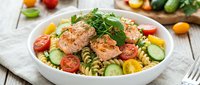 Grilled salmon pasta salad with fresh vegetables, cherry tomatoes, cucumber and herbs in white bowl on wooden table for healthy meal preparation.