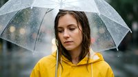 Young woman with long brown hair and fair complexion wearing bright yellow raincoat holding transparent umbrella in heavy rain, looking downward with serious expression, outdoor weather conditions.