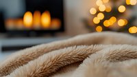 Cozy winter evening atmosphere with soft beige faux fur blanket in foreground and warm glowing bokeh lights from Christmas decorations in background.