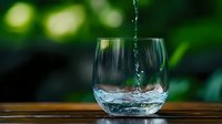 Clear water pouring into glass tumbler on wooden surface with green bokeh background. Fresh drinking water concept for health and hydration lifestyle.
