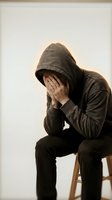 Depressed young man in hoodie covering face with hands sitting on chair expressing mental health struggles and emotional pain.